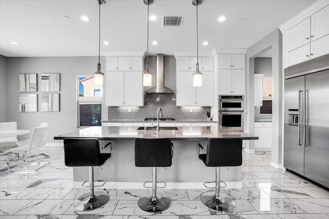 kitchen with pendant lighting, appliances with stainless steel finishes, a kitchen island with sink, and wall chimney range hood