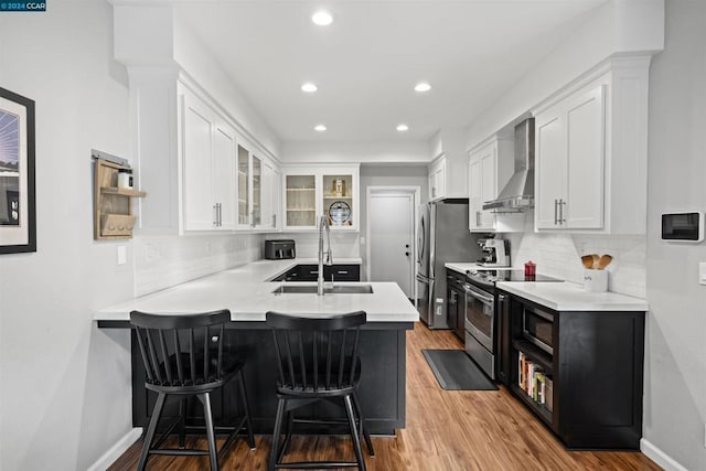 kitchen with wall chimney range hood, kitchen peninsula, sink, white cabinetry, and appliances with stainless steel finishes