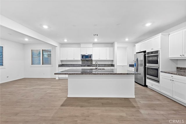 kitchen with stainless steel appliances, a kitchen island with sink, light hardwood / wood-style flooring, dark stone countertops, and white cabinets