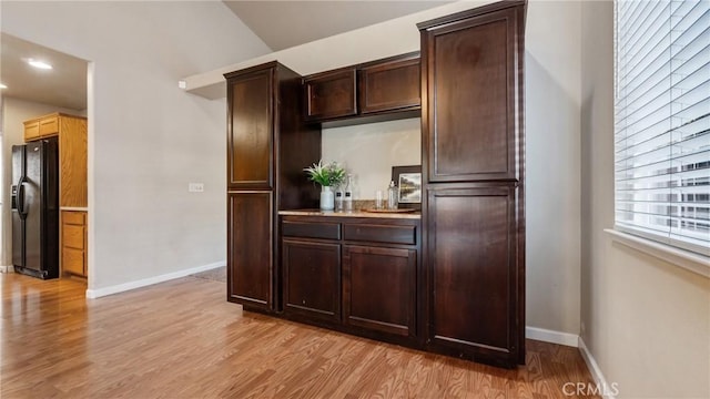 interior space featuring light wood-type flooring, black fridge with ice dispenser, vaulted ceiling, and dark brown cabinets