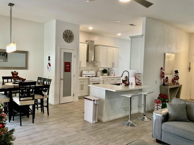 kitchen with white gas range oven, sink, wall chimney range hood, white cabinets, and hanging light fixtures