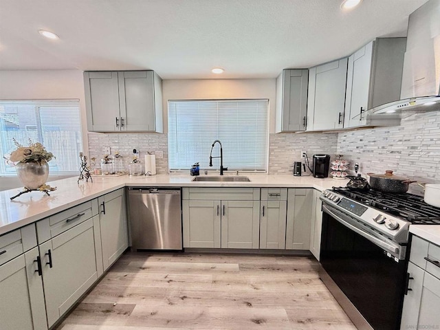 kitchen with gray cabinetry, sink, wall chimney range hood, light hardwood / wood-style flooring, and appliances with stainless steel finishes