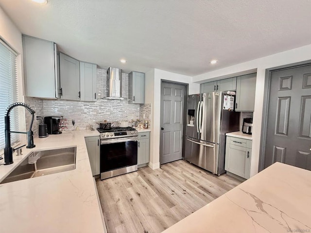 kitchen featuring light stone countertops, appliances with stainless steel finishes, wall chimney range hood, sink, and light hardwood / wood-style floors