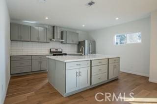 kitchen with dark wood-type flooring, stainless steel appliances, a kitchen island with sink, and wall chimney exhaust hood