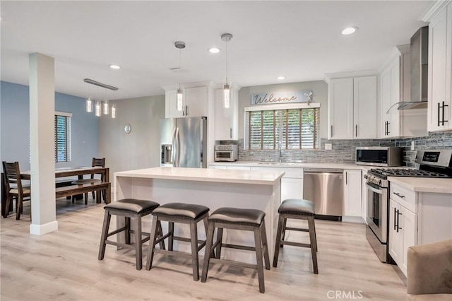 kitchen with wall chimney range hood, light wood-type flooring, decorative light fixtures, a kitchen island, and stainless steel appliances