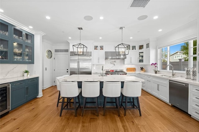 kitchen with appliances with stainless steel finishes, blue cabinetry, decorative light fixtures, light hardwood / wood-style flooring, and an island with sink