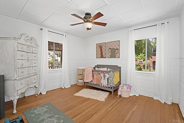 bedroom featuring multiple windows, ceiling fan, wood-type flooring, and a crib