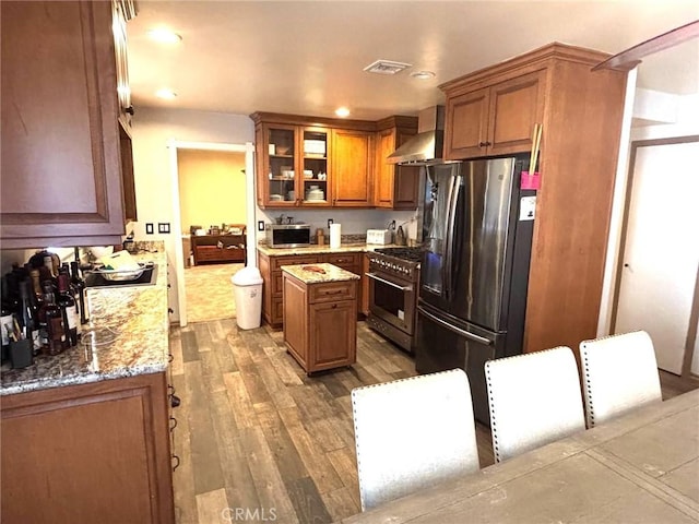 kitchen with appliances with stainless steel finishes, wood-type flooring, light stone countertops, wall chimney exhaust hood, and a center island