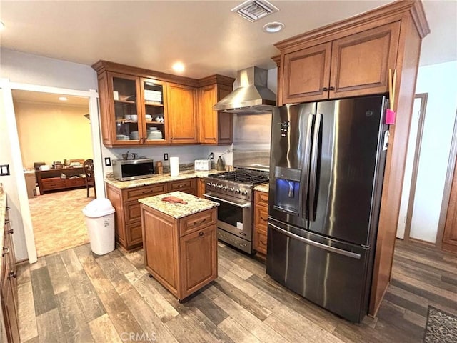 kitchen featuring hardwood / wood-style flooring, appliances with stainless steel finishes, light stone countertops, a kitchen island, and wall chimney range hood