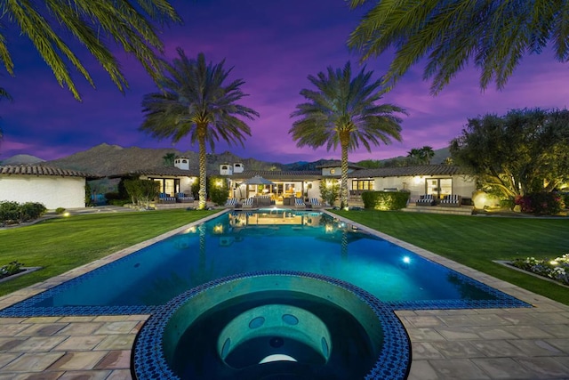 pool at dusk featuring a yard, an in ground hot tub, and a patio