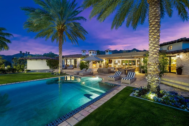 pool at dusk with a patio area, a yard, an outdoor bar, and an outdoor kitchen