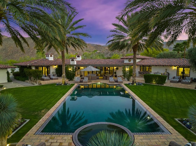 pool at dusk featuring a lawn, a patio area, and an in ground hot tub