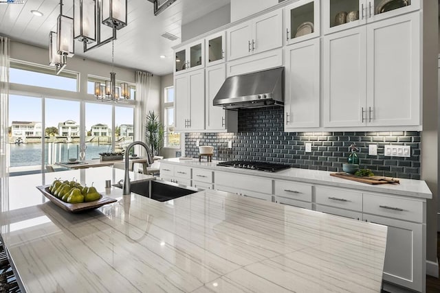 kitchen with sink, wall chimney range hood, light stone counters, pendant lighting, and a water view