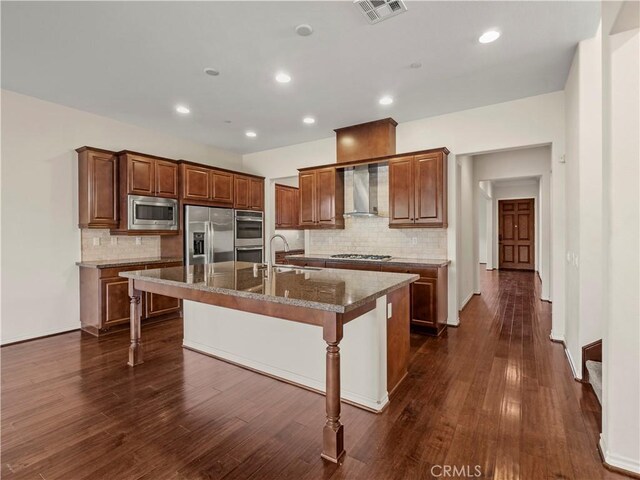 kitchen with wall chimney range hood, appliances with stainless steel finishes, a kitchen island with sink, dark stone countertops, and a kitchen bar