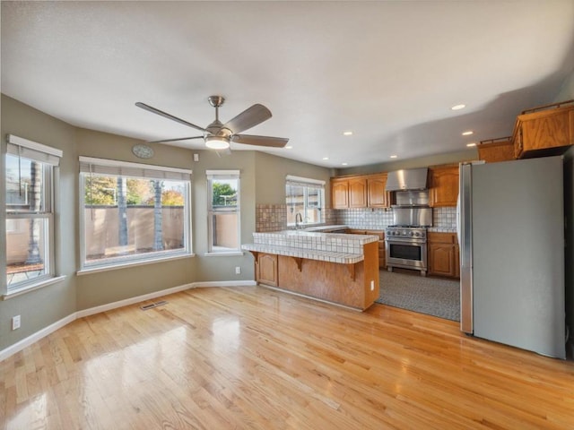 kitchen with wall chimney range hood, stainless steel appliances, kitchen peninsula, light wood-type flooring, and a breakfast bar area