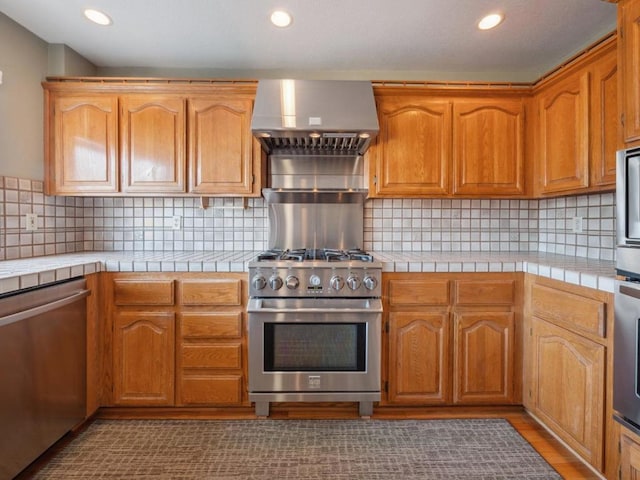 kitchen with backsplash, wall chimney range hood, stainless steel appliances, and tile countertops