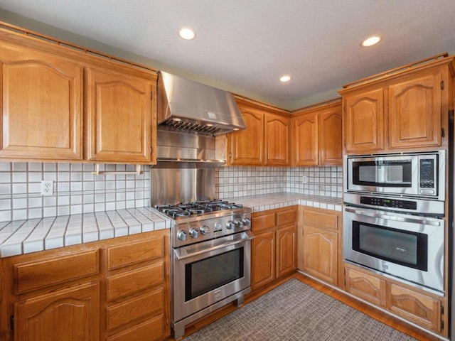 kitchen with wall chimney range hood, tile counters, appliances with stainless steel finishes, and tasteful backsplash