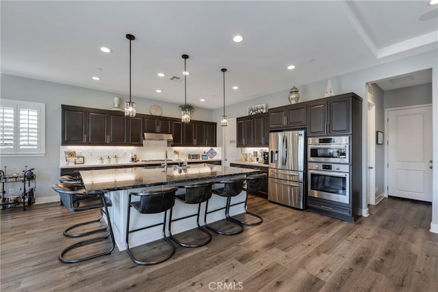 kitchen featuring dark hardwood / wood-style flooring, dark stone countertops, pendant lighting, a center island with sink, and appliances with stainless steel finishes