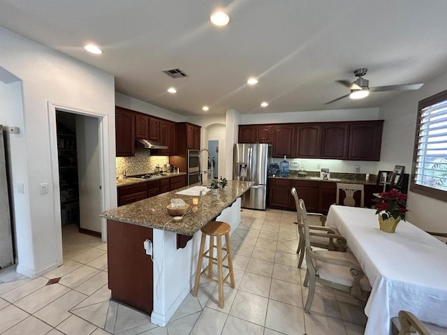 kitchen featuring stainless steel appliances, decorative backsplash, a kitchen breakfast bar, a kitchen island with sink, and ceiling fan