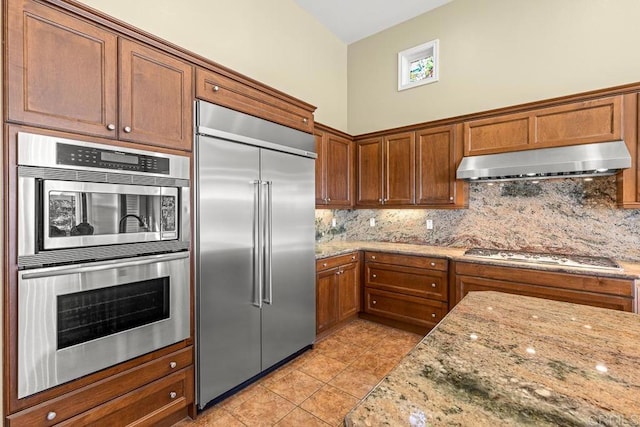 kitchen featuring appliances with stainless steel finishes, a towering ceiling, decorative backsplash, light stone counters, and light tile patterned floors