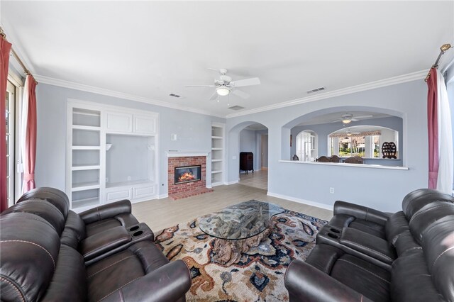 living room with built in shelves, ceiling fan, ornamental molding, and a brick fireplace