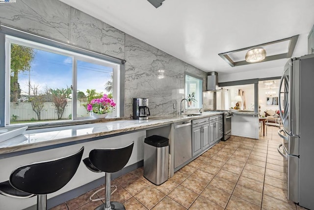 kitchen featuring appliances with stainless steel finishes, light stone counters, wall chimney exhaust hood, gray cabinetry, and sink