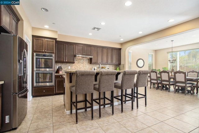 kitchen with pendant lighting, appliances with stainless steel finishes, an island with sink, backsplash, and a breakfast bar