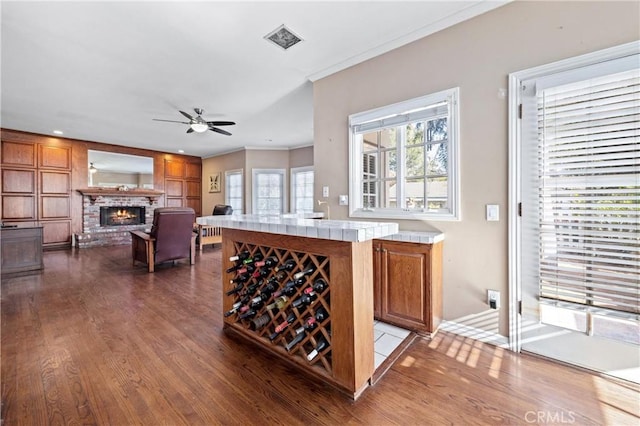 bar featuring dark hardwood / wood-style flooring, ceiling fan, tile countertops, a brick fireplace, and crown molding