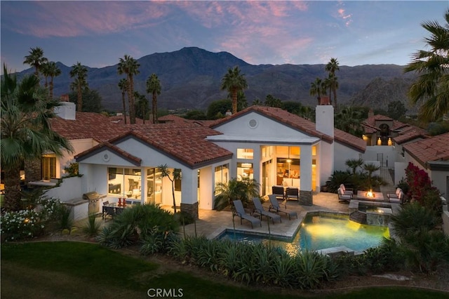 back house at dusk featuring a mountain view, an outdoor fire pit, a pool with hot tub, area for grilling, and a patio