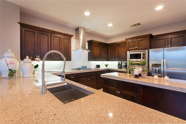 kitchen with light stone countertops, appliances with stainless steel finishes, wall chimney exhaust hood, dark brown cabinetry, and sink