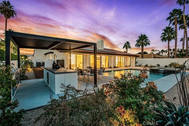 back house at dusk featuring area for grilling, a swimming pool with hot tub, an outdoor bar, and a patio