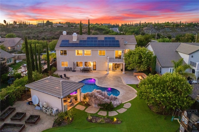 back house at dusk featuring solar panels, a patio, a balcony, and a lawn