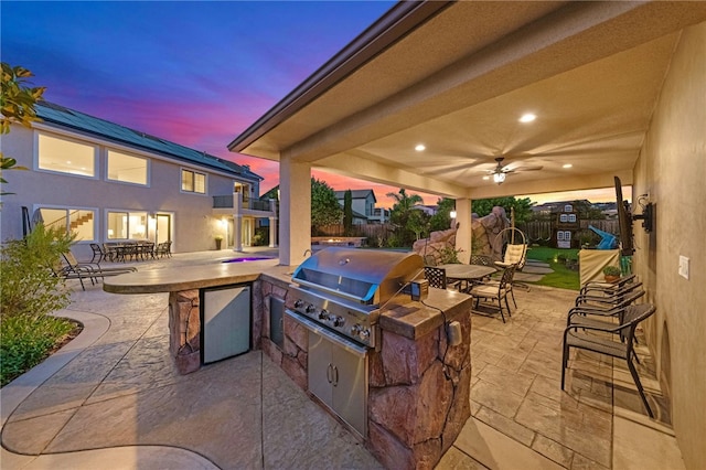 patio terrace at dusk featuring area for grilling, ceiling fan, exterior bar, and exterior kitchen