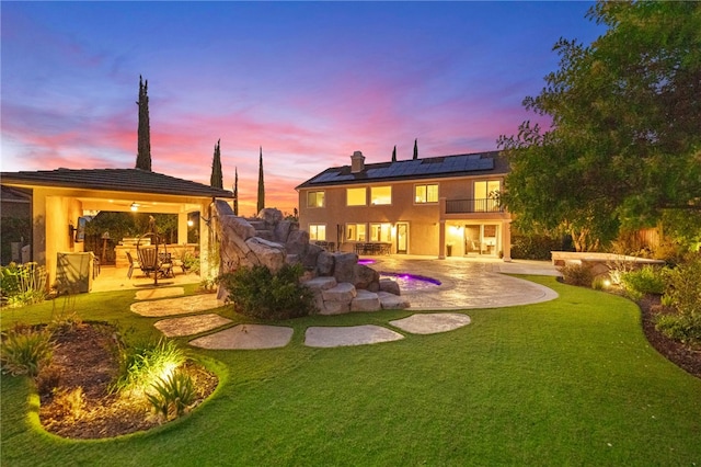 back house at dusk featuring a lawn, a patio area, a balcony, and solar panels