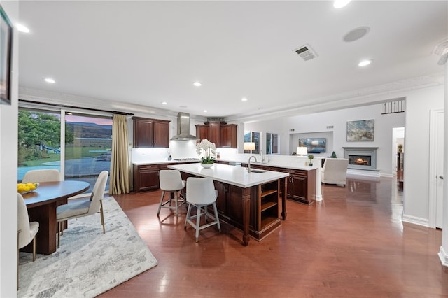 kitchen featuring wall chimney exhaust hood, dark brown cabinets, dark wood-type flooring, a kitchen island, and a breakfast bar area