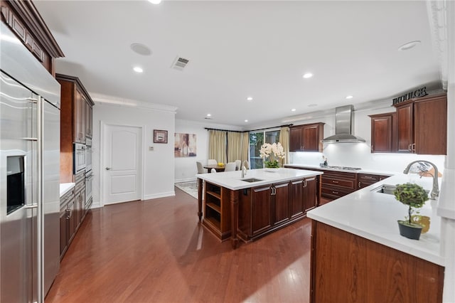 kitchen featuring dark wood-type flooring, wall chimney range hood, sink, an island with sink, and stainless steel appliances