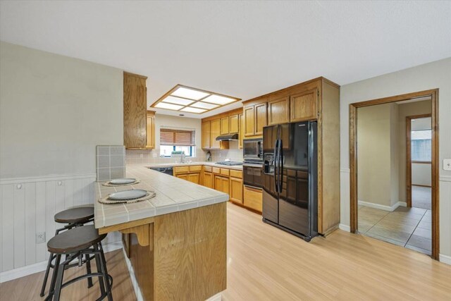 kitchen featuring black appliances, kitchen peninsula, light wood-type flooring, and tile countertops