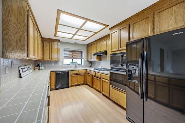 kitchen with tile countertops, tasteful backsplash, light wood-type flooring, black appliances, and sink