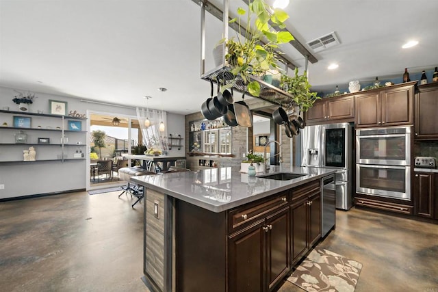 kitchen with a center island with sink, sink, stainless steel appliances, hanging light fixtures, and dark brown cabinets