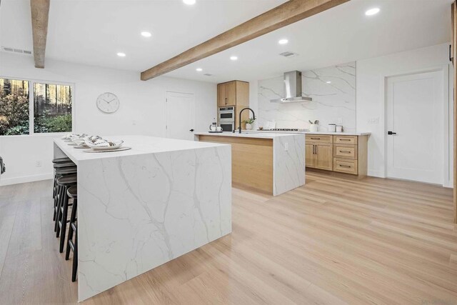 kitchen with a breakfast bar, wall chimney range hood, light hardwood / wood-style flooring, beamed ceiling, and an island with sink