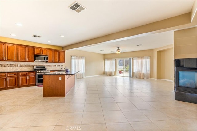 kitchen featuring light tile patterned floors, a center island with sink, ceiling fan, appliances with stainless steel finishes, and decorative backsplash