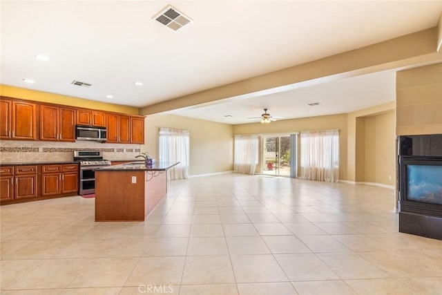 kitchen featuring appliances with stainless steel finishes, backsplash, a kitchen island with sink, ceiling fan, and a tiled fireplace