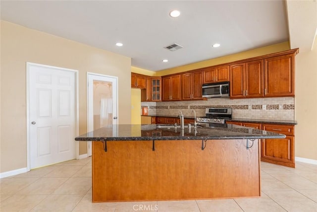 kitchen with sink, light tile patterned flooring, appliances with stainless steel finishes, an island with sink, and dark stone counters