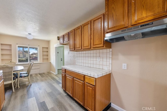 kitchen with under cabinet range hood, light wood-type flooring, brown cabinets, and baseboards