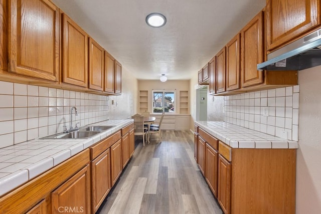 kitchen featuring under cabinet range hood, tile countertops, light wood-style flooring, brown cabinets, and a sink