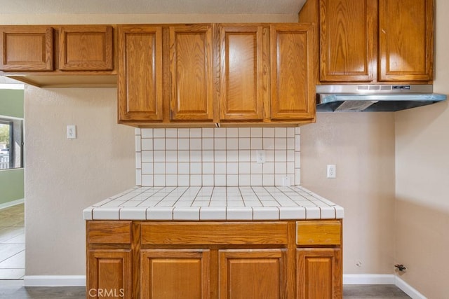 kitchen featuring under cabinet range hood, brown cabinets, tasteful backsplash, and baseboards