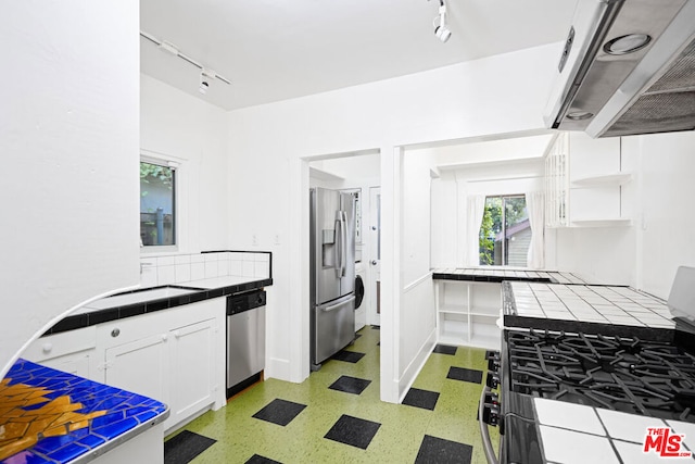 kitchen with track lighting, appliances with stainless steel finishes, tile counters, white cabinetry, and extractor fan