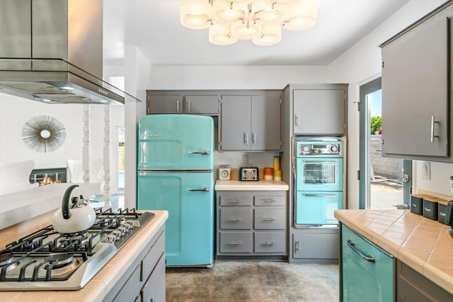 kitchen with stainless steel gas stovetop, double wall oven, wall chimney exhaust hood, tile counters, and white fridge