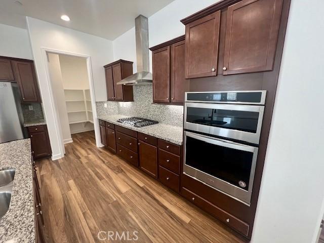 kitchen featuring light stone countertops, dark brown cabinetry, wall chimney exhaust hood, stainless steel appliances, and light hardwood / wood-style flooring