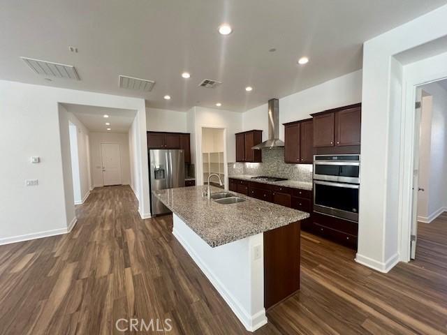 kitchen with a center island with sink, appliances with stainless steel finishes, wall chimney range hood, light stone countertops, and sink